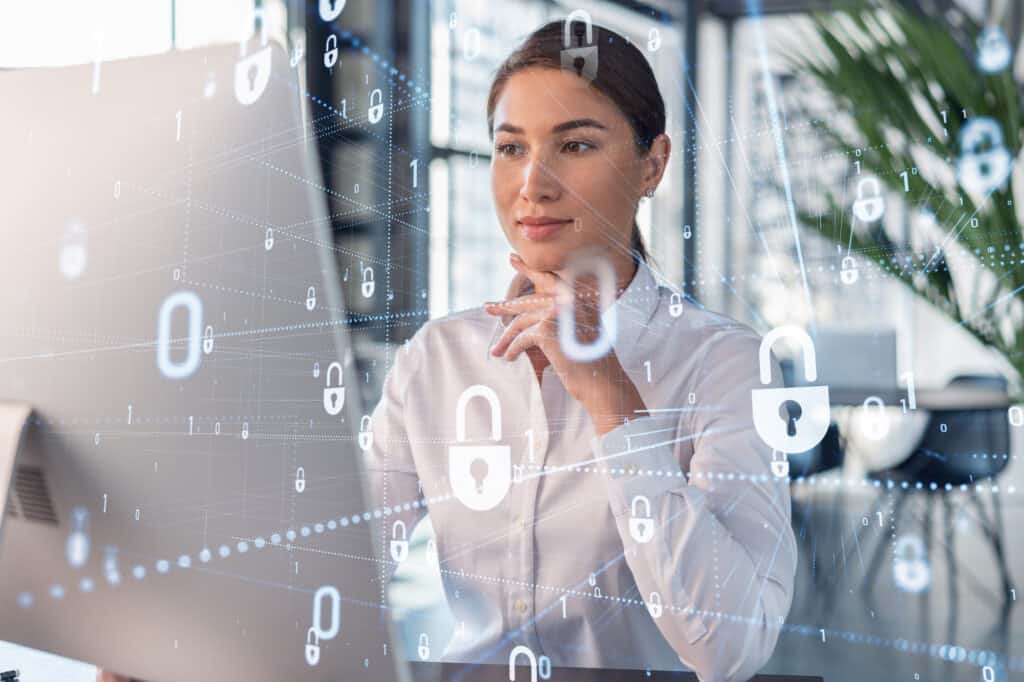 Attractive businesswoman in white shirt at workplace working with laptop to defend customer cyber security. Concept of clients information protection. Padlock hologram over office background.